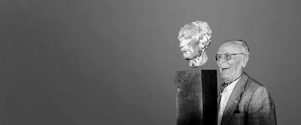 Historische Schwarz-Weiß-Fotografie eines lächelnden Friedrich Schiedel, der neben einer auf einem dunklen Sockel montierten Porträtbüste steht.