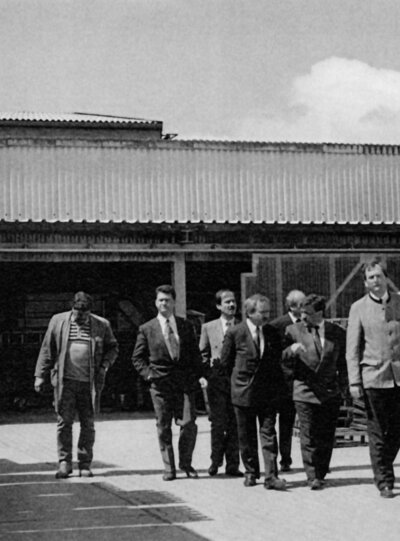 Eine historische Schwarz-Weiß-Fotografie, die eine Gruppe von Männern in Business-Anzügen zeigt, die durch eine Schiedel Industrieanlage im Freien gehen und dabei an gestapelten Holzpaletten und verpackten Schiedel-Produkten vorbeikommen.