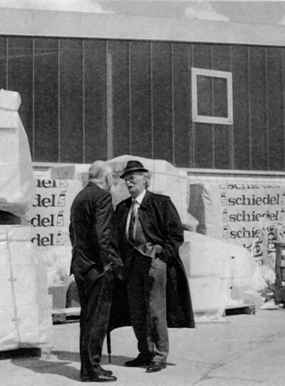 Eine historische Schwarz-Weiß-Fotografie von zwei Männern in Mänteln, die sich außerhalb einer Schiedel-Produktionsstätte neben gestapelten Paletten mit verpackten Produkten unterhalten.