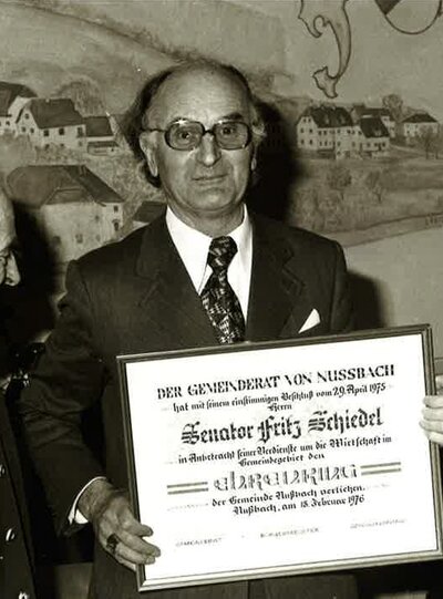 Eine historische Schwarz-Weiß-Fotografie von Friedrich Schiedel, der stolz eine offizielle, gerahmte Ehrenurkunde hält, flankiert von zwei weiteren Männern in Anzügen.