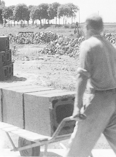 Eine historische Schwarz-Weiß-Fotografie, die einen Arbeiter von hinten zeigt, der eine mit schweren Schiedel-Mantelsteinen beladene Metallschubkarre über das Außengelände einer Fabrik schiebt.