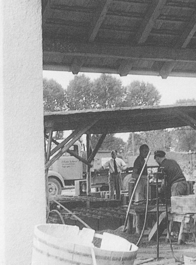 Eine historische Schwarz-Weiß-Fotografie von Arbeitern, die Maschinen unter einem hölzernen Freiluftunterstand in einer frühen Schiedel-Betriebsstätte bedienen, im Hintergrund ist ein historischer Lieferwagen zu sehen.