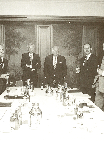 Eine historische Schwarz-Weiß-Fotografie von Friedrich Schiedel im formellen Anzug und mit Fliege, der während einer feierlichen Zusammenkunft an einem langen Konferenztisch spricht, während Führungskräfte die Weingläser heben, um einen Meilenstein des Unternehmens zu feiern.